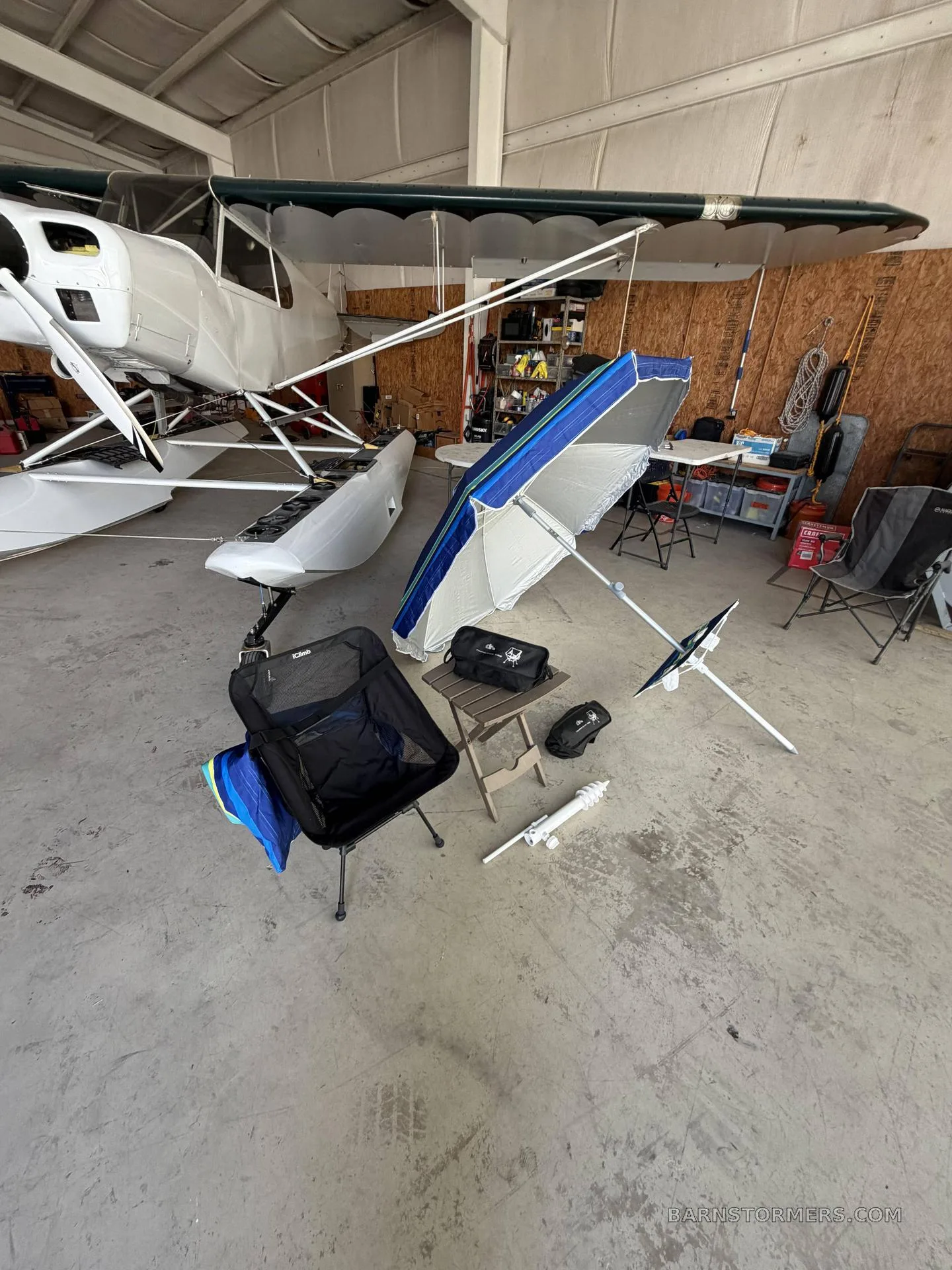 A white seaplane with floats is parked inside a hangar, with camping chairs, a table, and an umbrella set up nearby.