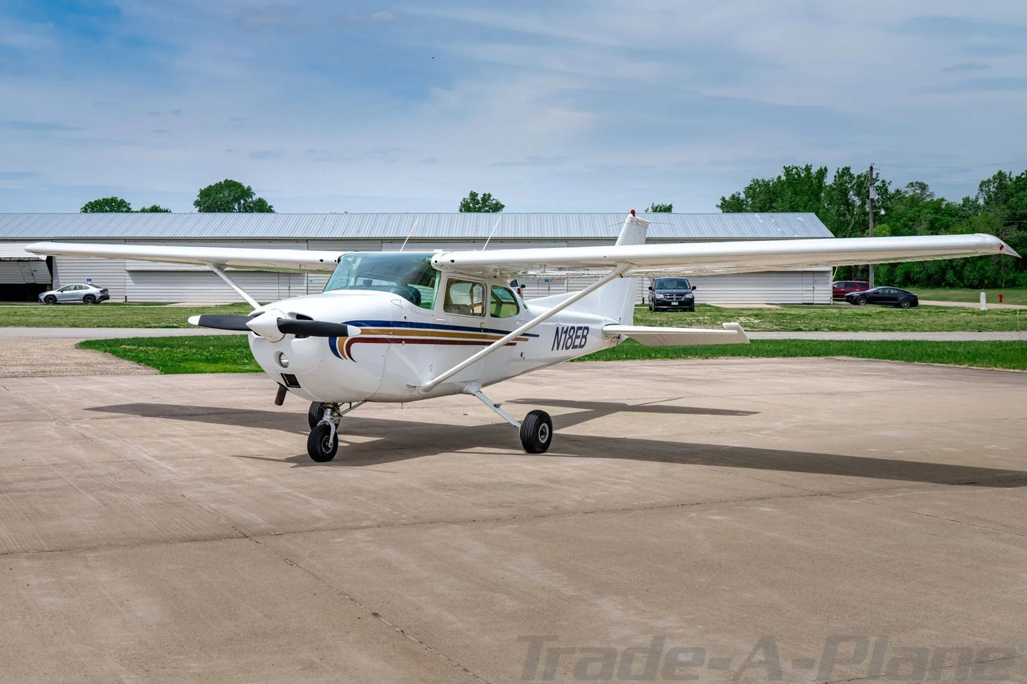 A white Cessna 172 with blue, gold, and red stripes on the nose and fuselage, parked on an asphalt ramp with buildings in the background.