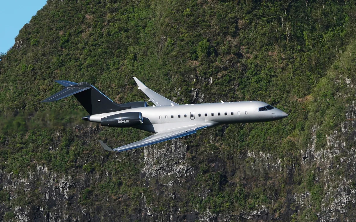 A silver and black Bombardier Global 6000 business jet with registration 9H-ARE flies past a lush green, rocky mountain.