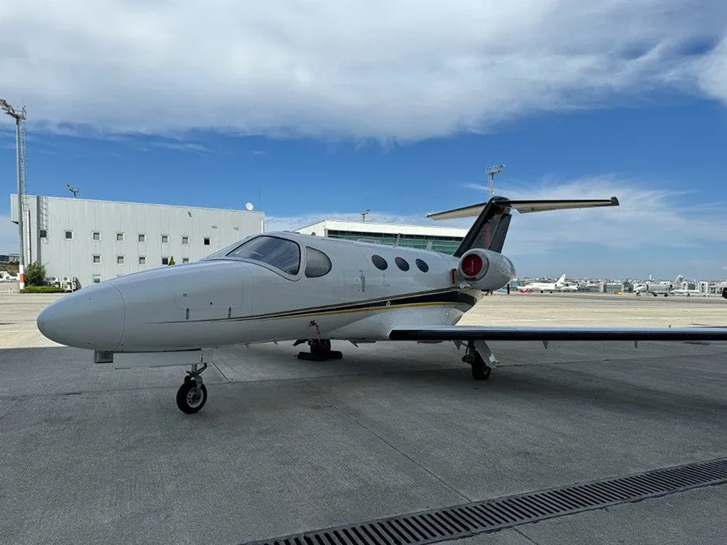A white business jet with black and gold stripes on the tail, parked on an airport tarmac with buildings and other aircraft in the background.