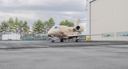 A beige and gold private jet with a unique swirl paint scheme is parked on an asphalt apron next to a white hangar.