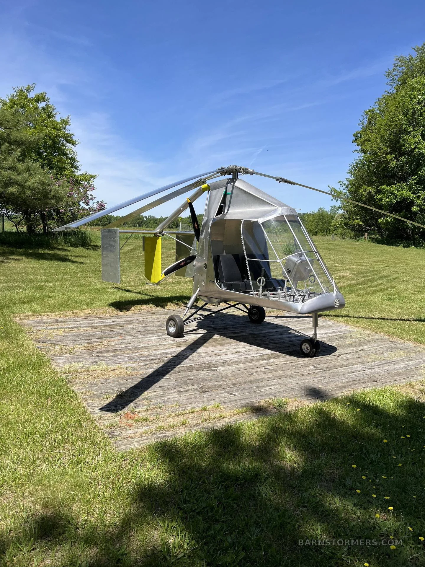 A silver single-seat ultralight helicopter/autogyro with a yellow tail fin, parked on a wooden pad in a grassy field under a clear blue sky.
