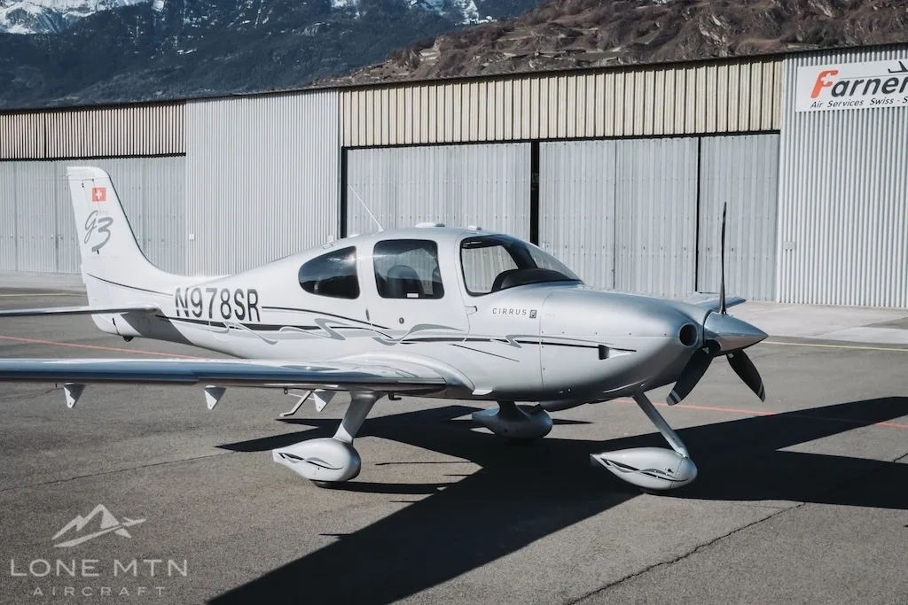 A silver Cirrus SR22 aircraft (N978SR) parked on an airfield next to a hangar.