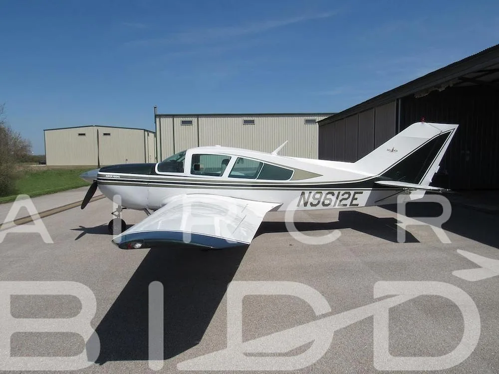 A side view of the white and dark green single-engine aircraft N9612E parked on a tarmac with hangars in the background.