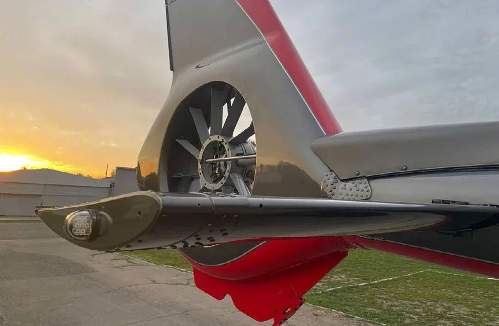 A close-up view of the fenestron (enclosed tail rotor) of a grey and red helicopter at sunset.