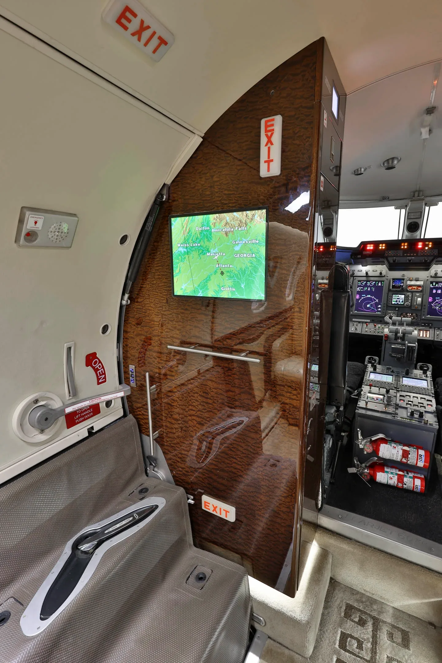 The aircraft cabin entry area with a wood-paneled wall, a screen displaying a map, and a partial view of the cockpit.