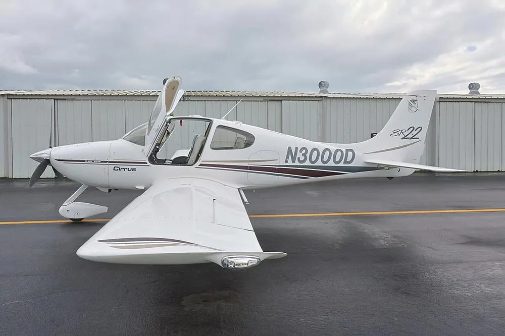 Left side view of a white Cirrus SR22 aircraft with N3000D registration and the pilot door open, parked on a wet tarmac.