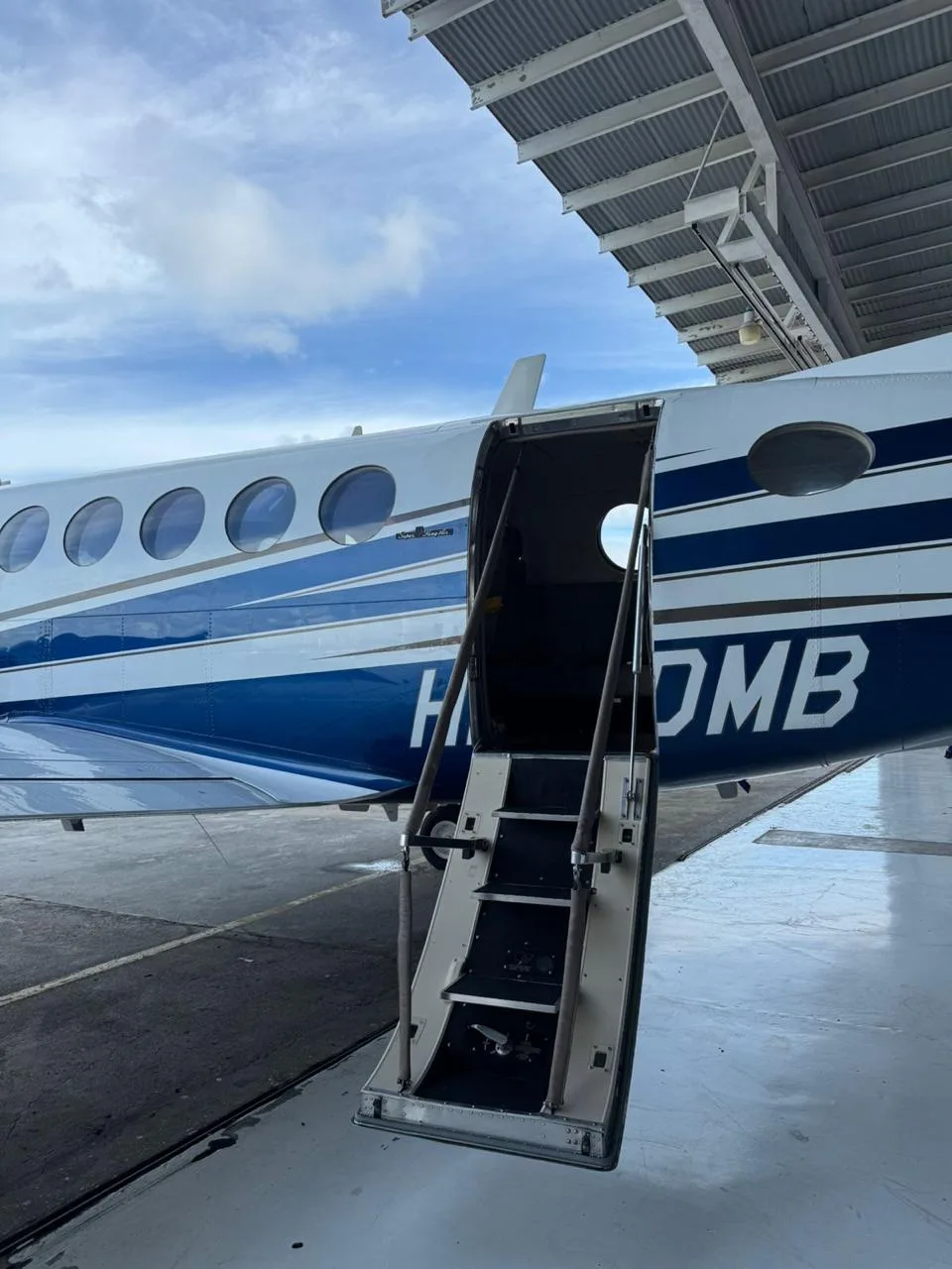 The open airstair door of a white and blue turboprop aircraft (King Air) with registration HP-5DMB, parked under a hangar.