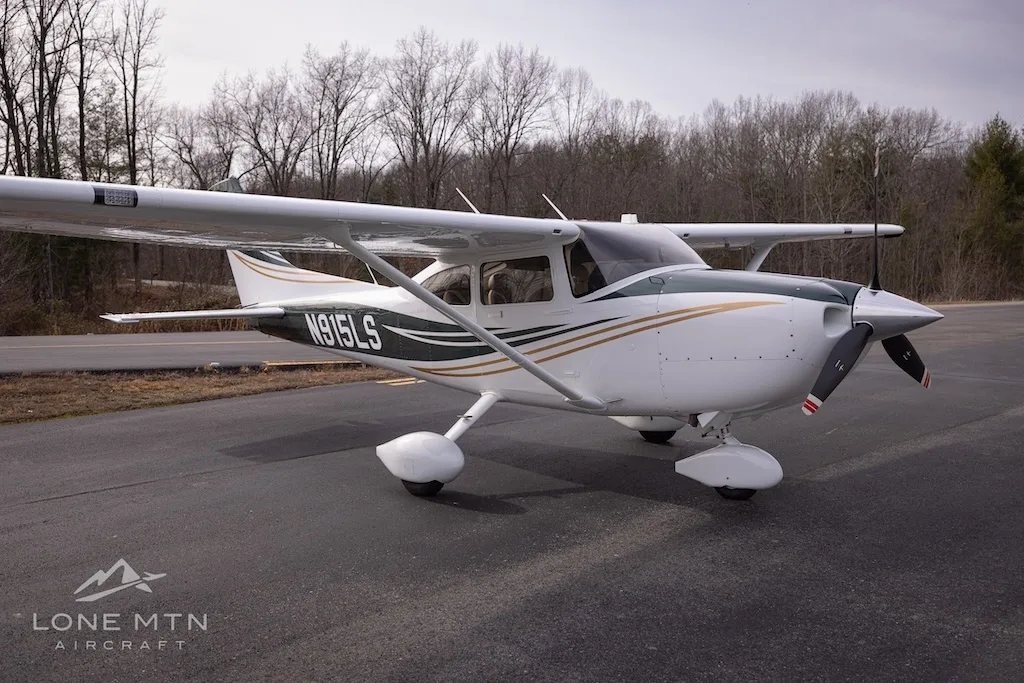 A white and dark green Cessna Skylane N915LS with gold stripes, parked on an asphalt ramp with trees in the background.