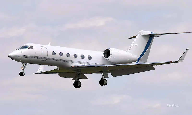 A white Gulfstream business jet with a blue stripe on its tail descends with landing gear extended against a cloudy sky.
