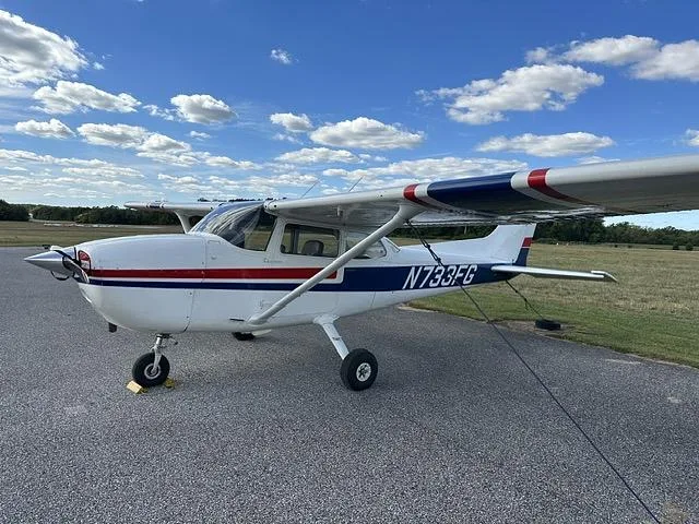 A white Cessna 172 Skyhawk with blue and red stripes, registration N733FG, parked on an asphalt ramp under a blue sky with clouds.