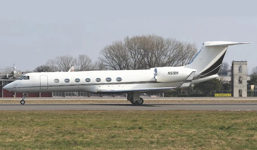 A white Gulfstream G550 business jet with black and gold accents on the tail, taxiing on a runway with trees and buildings in the background.