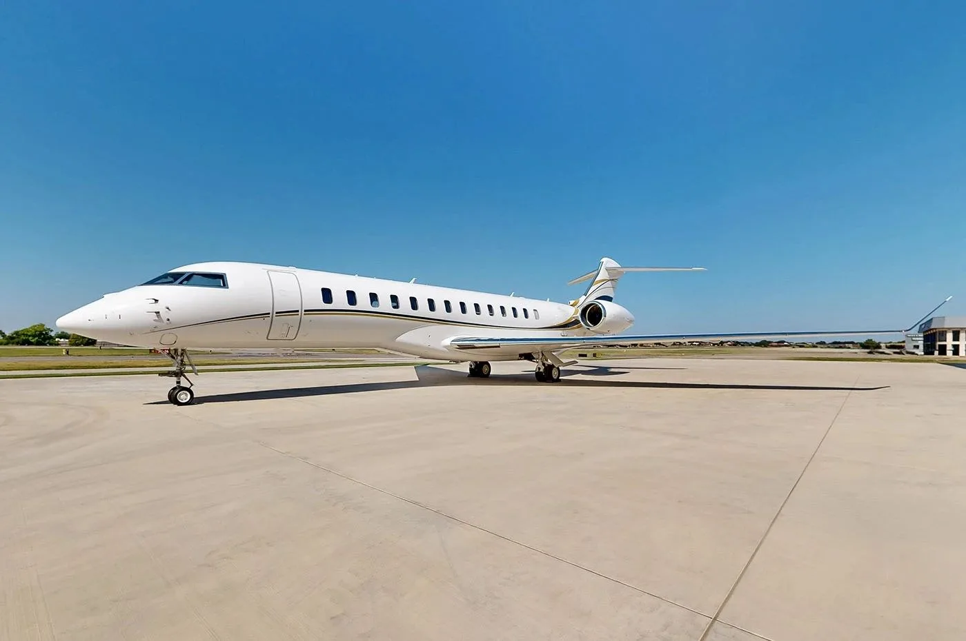 A white Bombardier Global business jet with gold and black stripes parked on a tarmac under a clear blue sky.