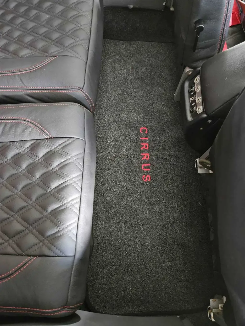 A dark grey floor mat with "CIRRUS" embroidered in red, between two black leather aircraft seats.