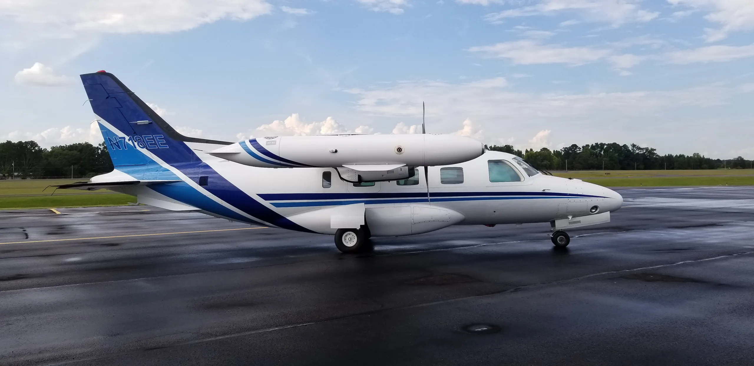 A white twin-engine turboprop aircraft with blue stripes on the tail and fuselage, registered N718EE, parked on a wet tarmac.