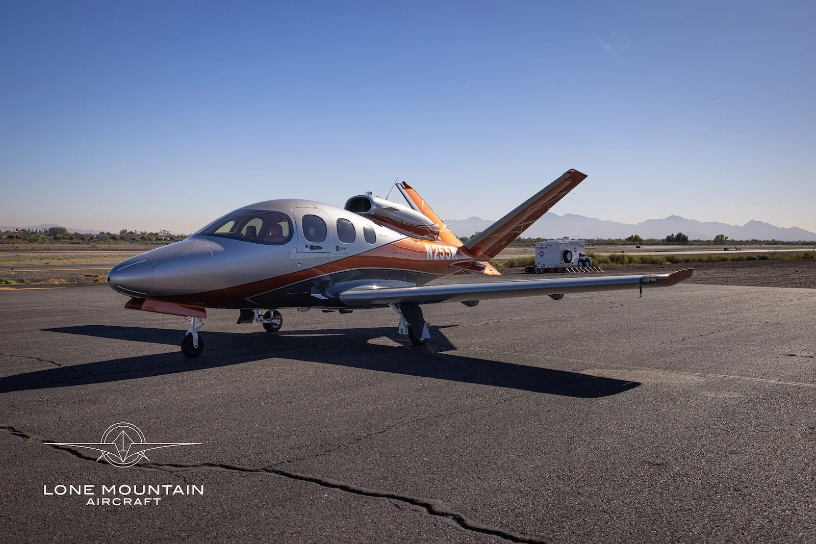 A three-quarter front view of a silver and orange Cirrus Vision Jet (SF50) parked on an asphalt tarmac with mountains in the distance.