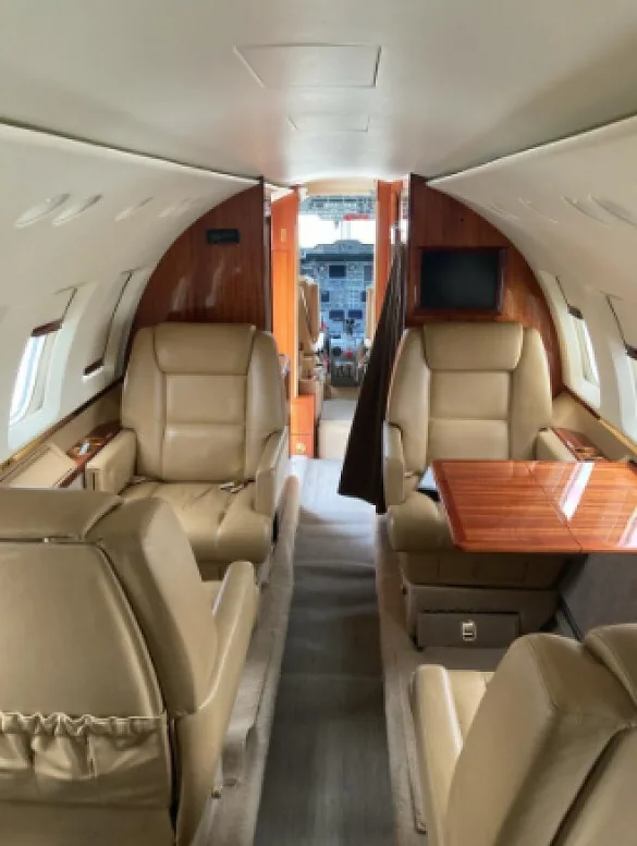 The interior cabin of an aircraft, featuring tan leather seats, wooden tables, and an aisle leading to the cockpit.