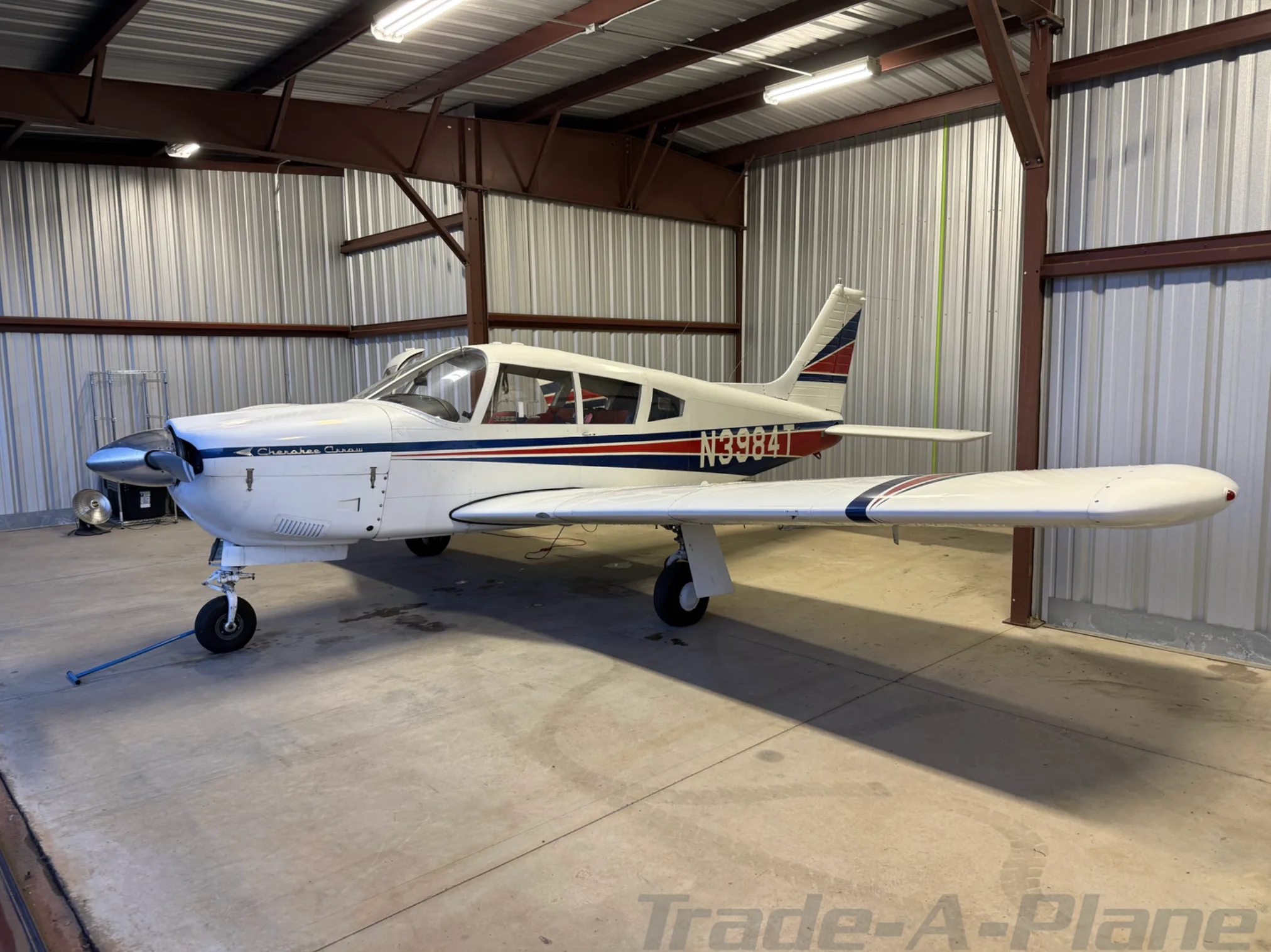 A white Piper Cherokee Arrow aircraft with blue and red stripes, registration N3984T, parked inside a hangar.