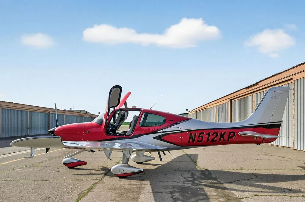 A red and silver single-engine aircraft, N512KP, with its gull-wing doors open, parked on an airfield next to hangars.