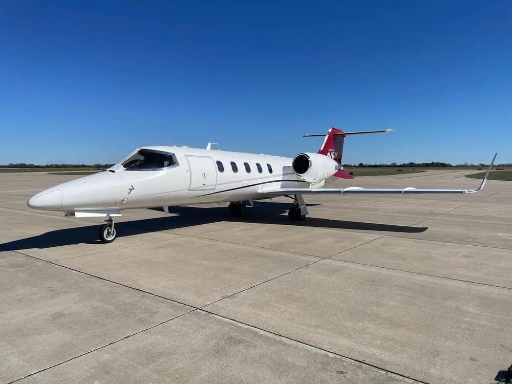 A white Learjet with red tail and accents, registration N70AY, parked on an airport tarmac under a clear blue sky.