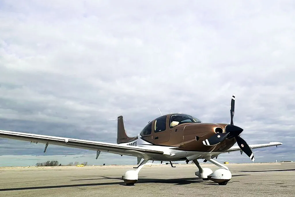 A Cirrus SR22 aircraft with a brown and white paint scheme parked on an airfield under a cloudy sky.