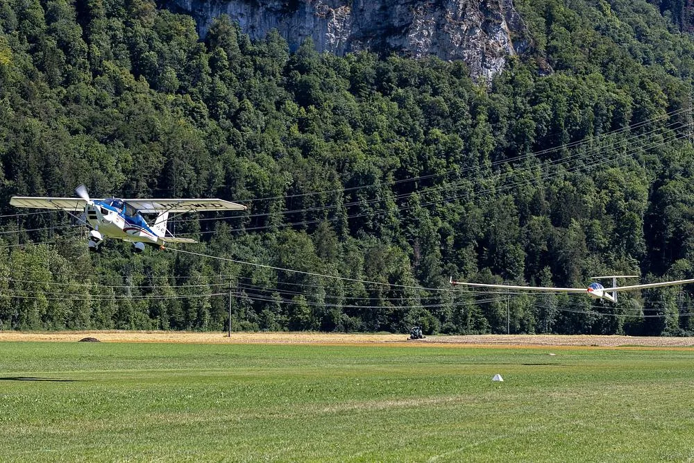 A white and blue tow plane takes off, pulling a white glider over a green field with a forested mountain in the background.
