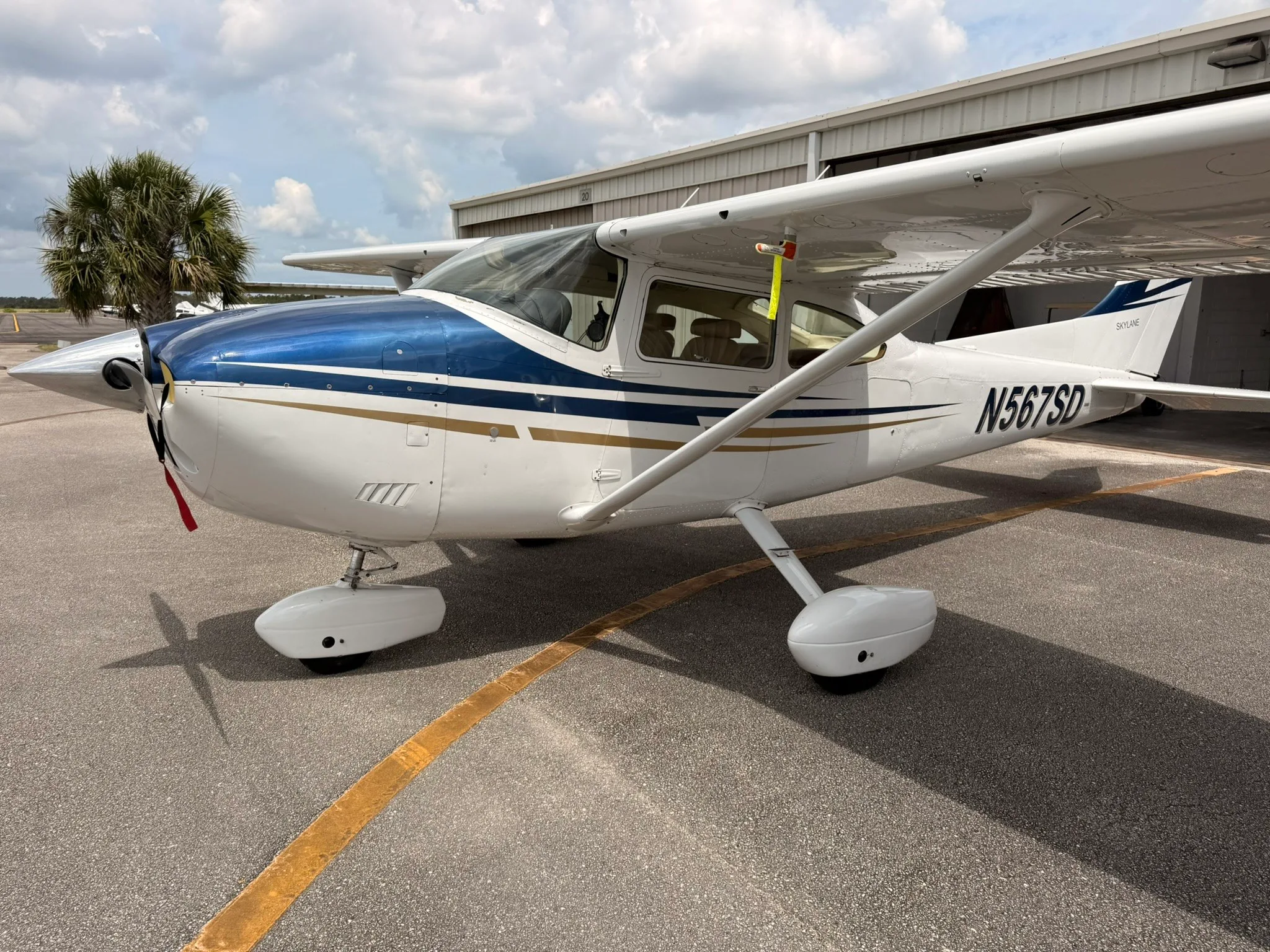 A front-left view of a white Cessna Skylane N567SD with blue and gold stripes parked on an asphalt ramp.