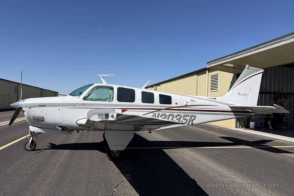A white Beechcraft Baron with maroon and grey stripes, registration N2035R, parked on an asphalt ramp next to a hangar.