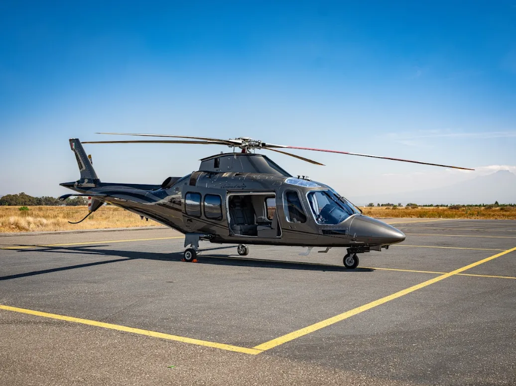 A grey AgustaWestland AW109 GrandNew helicopter with an open passenger door is parked on an airfield under a clear blue sky.