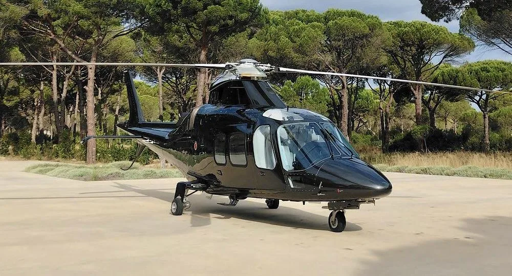 A dark grey helicopter with its main rotor blades extended, parked on a concrete pad surrounded by tall pine trees.