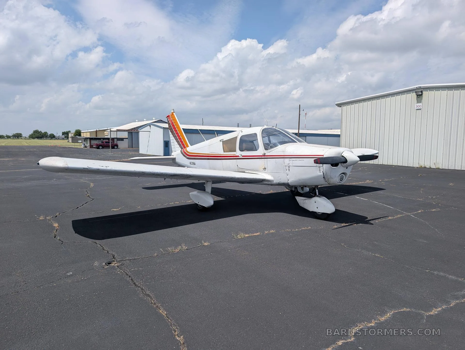 A white single-engine low-wing aircraft with red, orange, and brown stripes on the tail and fuselage, parked on an asphalt ramp with hangars in the background.
