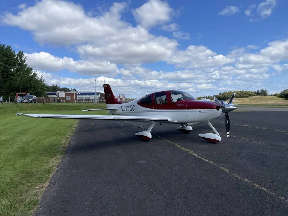 A red and white Cirrus SR22 aircraft, registration N925CC, is parked on an asphalt ramp under a partly cloudy sky.