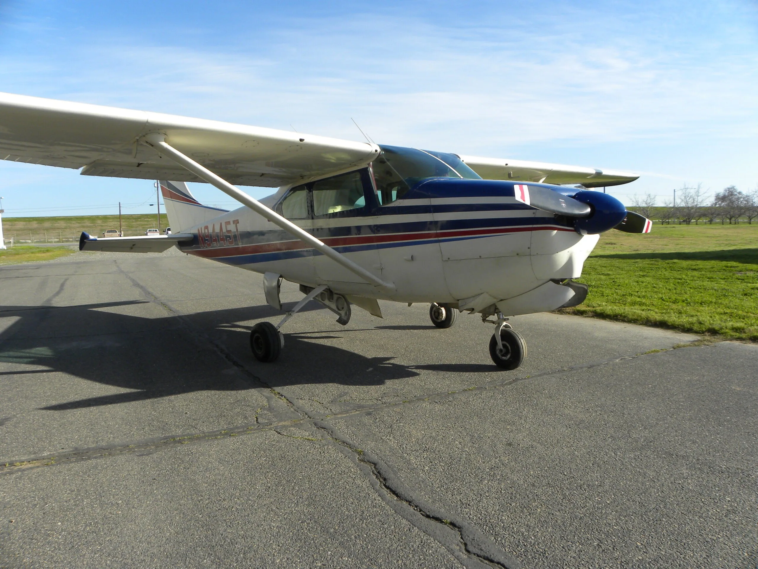 A white Cessna 182 with blue and red stripes parked on a tarmac with grass in the background.