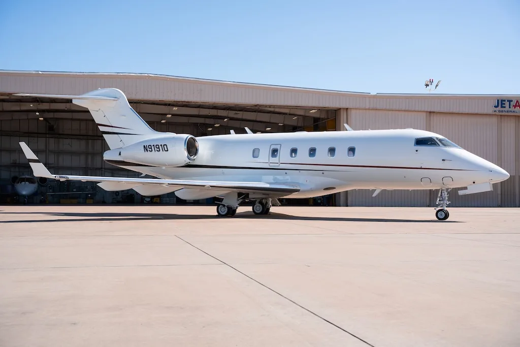 A white business jet with a red stripe is parked on a tarmac in front of a hangar under a clear blue sky.