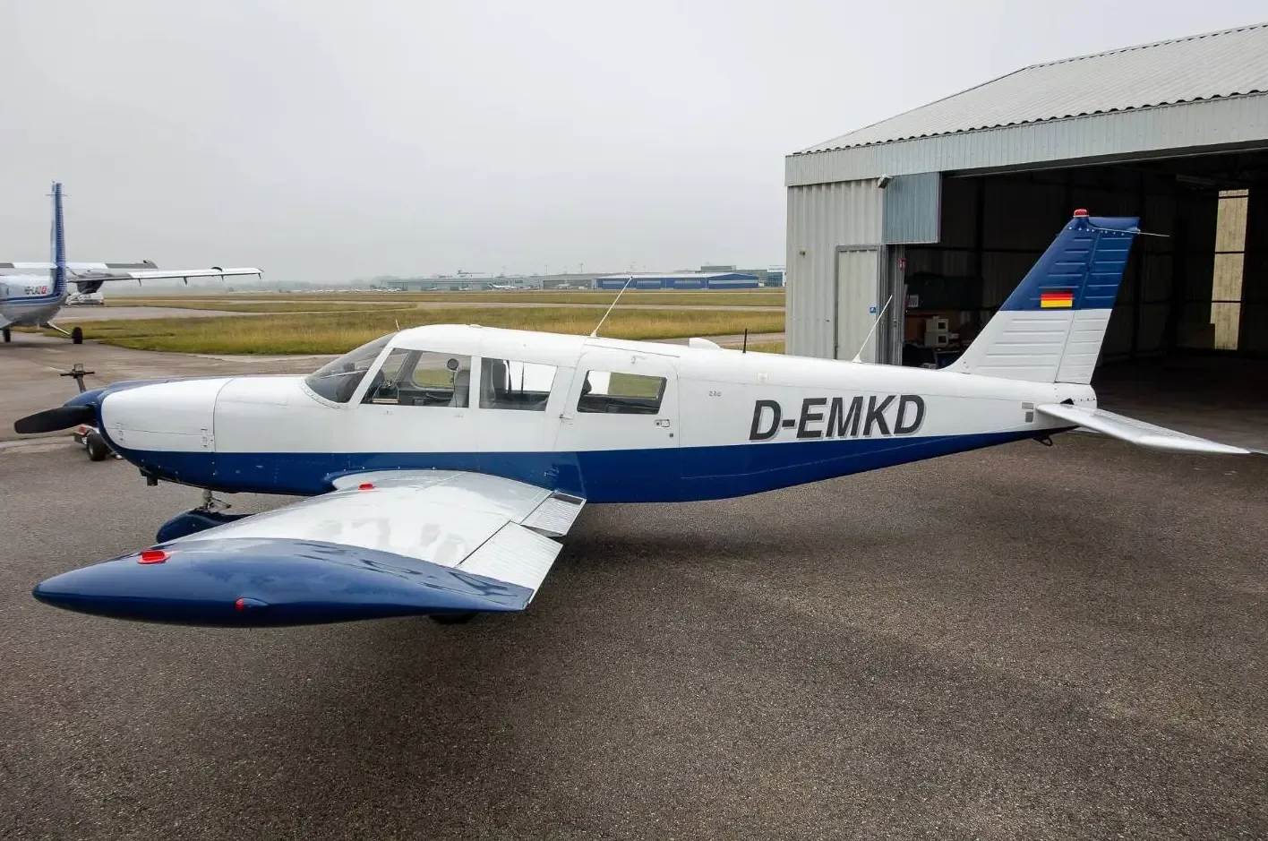 A white and blue single-engine propeller aircraft (D-EMKD) with a German flag on its tail, parked on an asphalt apron.