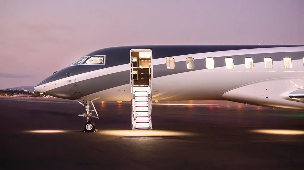 A close-up side view of a grey and white private jet, likely a Global 7500, with its illuminated main door open and stairs extended on a tarmac at dusk.