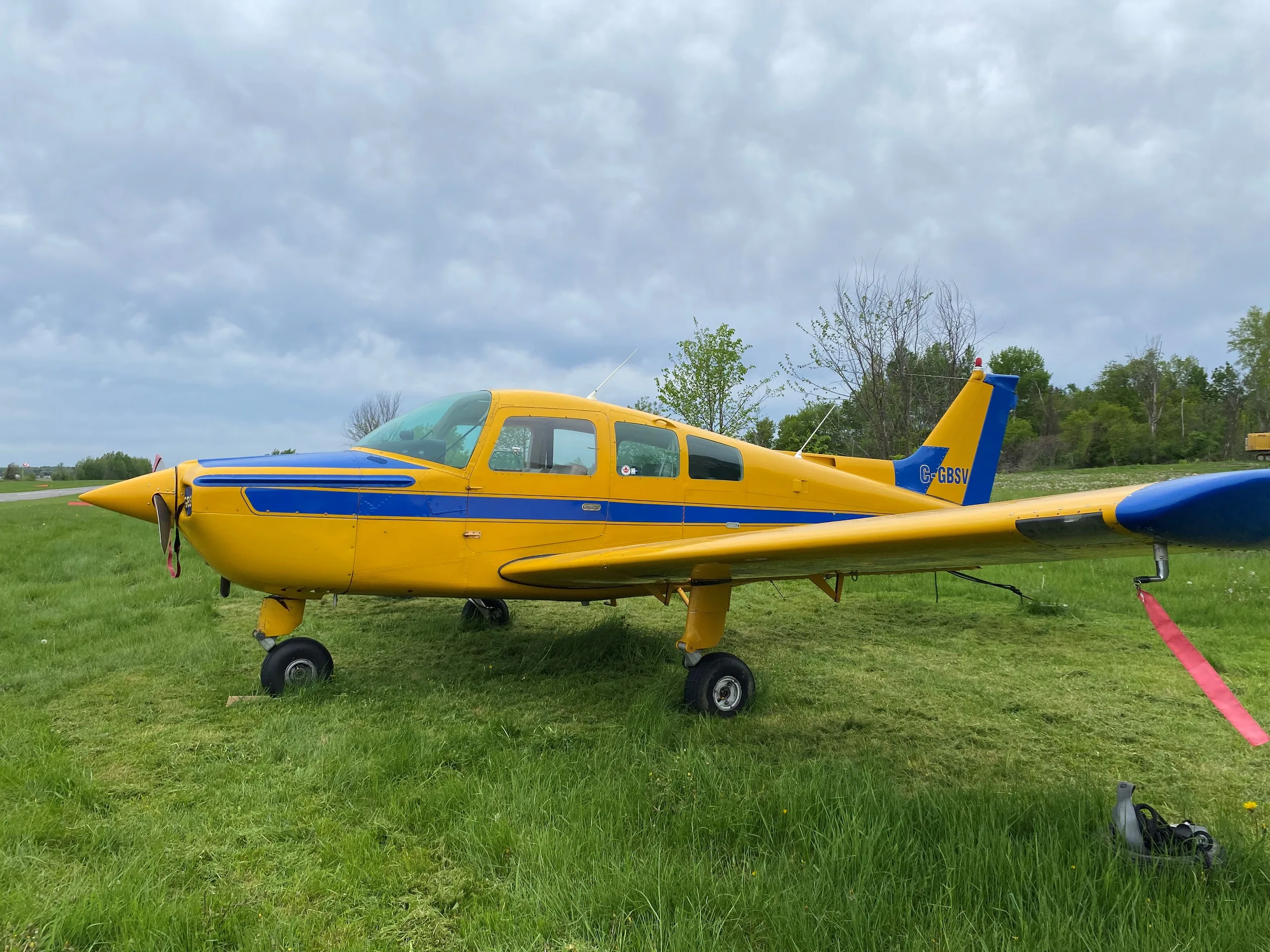 A yellow and blue single-engine aircraft with registration C-GBSV parked on a grassy field under a cloudy sky.