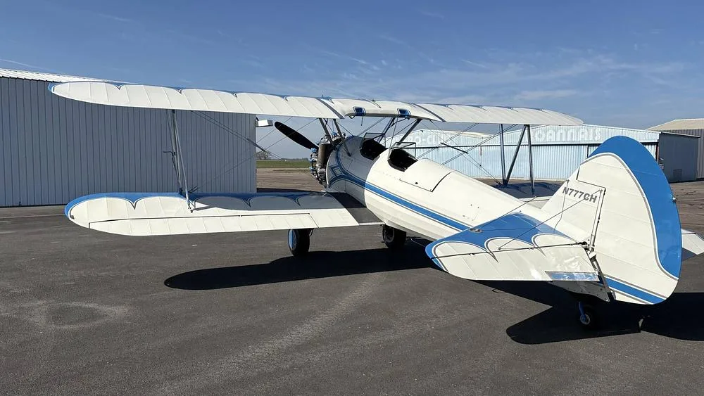 A white and blue biplane, N777CH, parked on an airfield with hangars in the background.