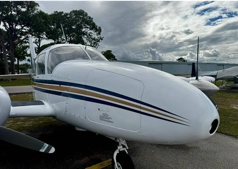 A front-side view of the white nose and fuselage of a twin-engine aircraft with blue and gold stripes, parked on an airfield.