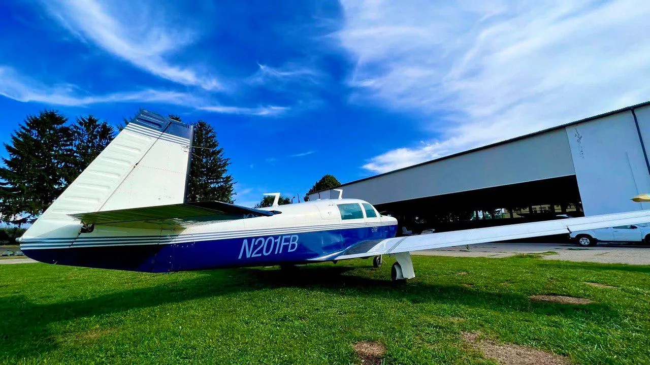 A rear-left view of the N201FB single-engine aircraft parked on grass under a bright blue sky.