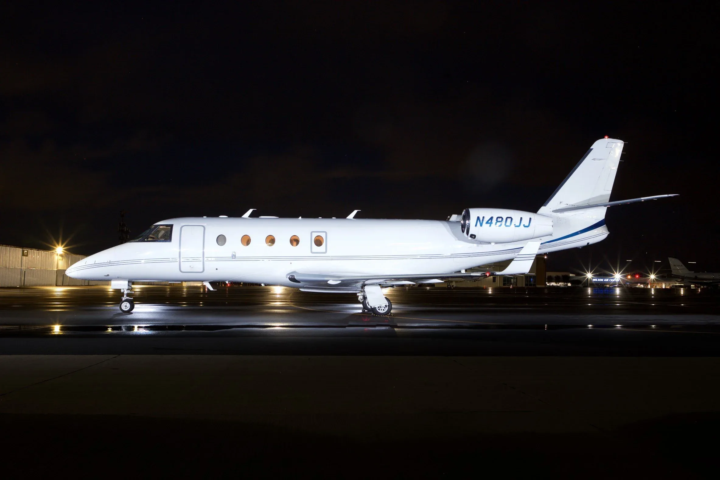 A white business jet with blue tail stripes is parked on a wet tarmac at night, illuminated by ground lights.