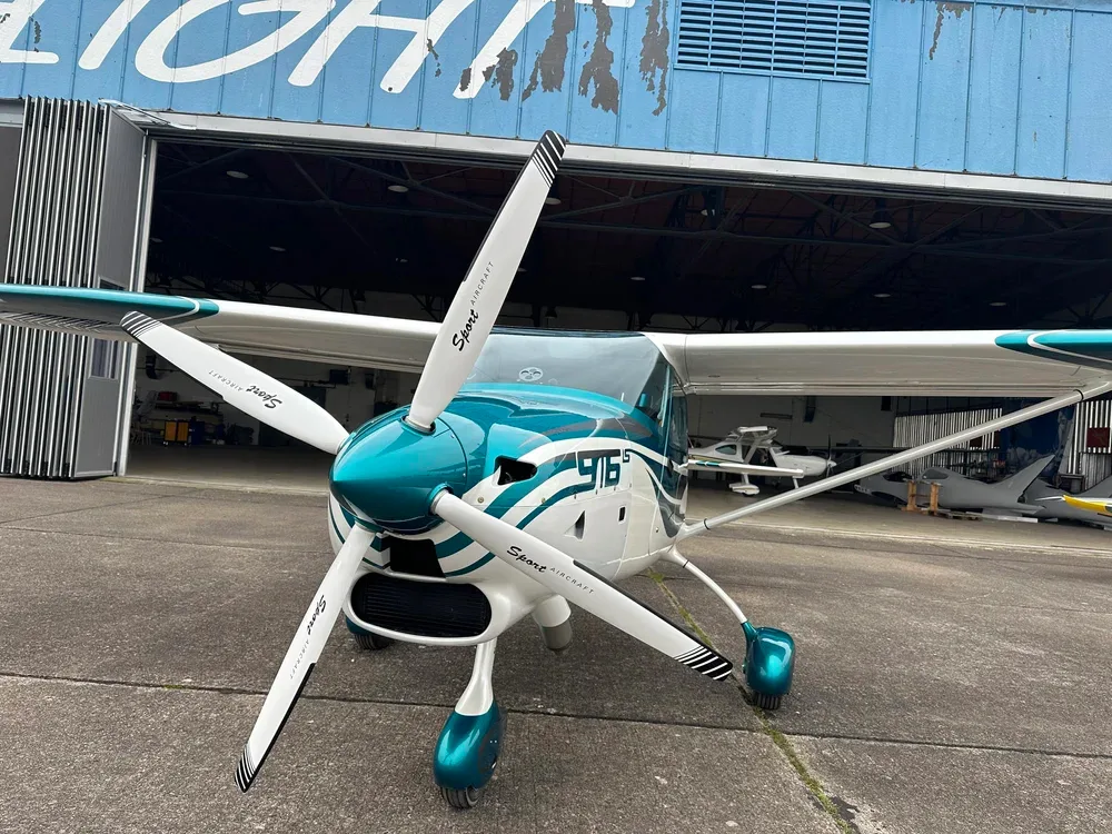 A head-on view of a white and teal Sirius 916 LS aircraft, registration N84TL, with its propeller prominently displayed, parked in front of a hangar.