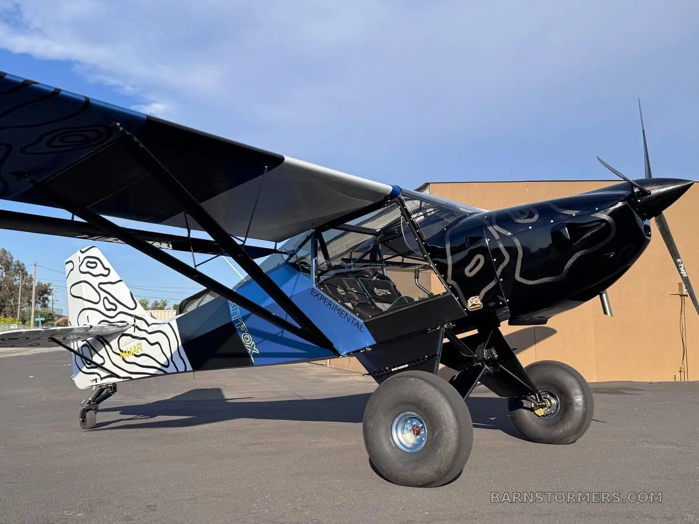 A black and blue Kitfox aircraft with "EXPERIMENTAL" text and large tundra tires, parked on an asphalt tarmac.