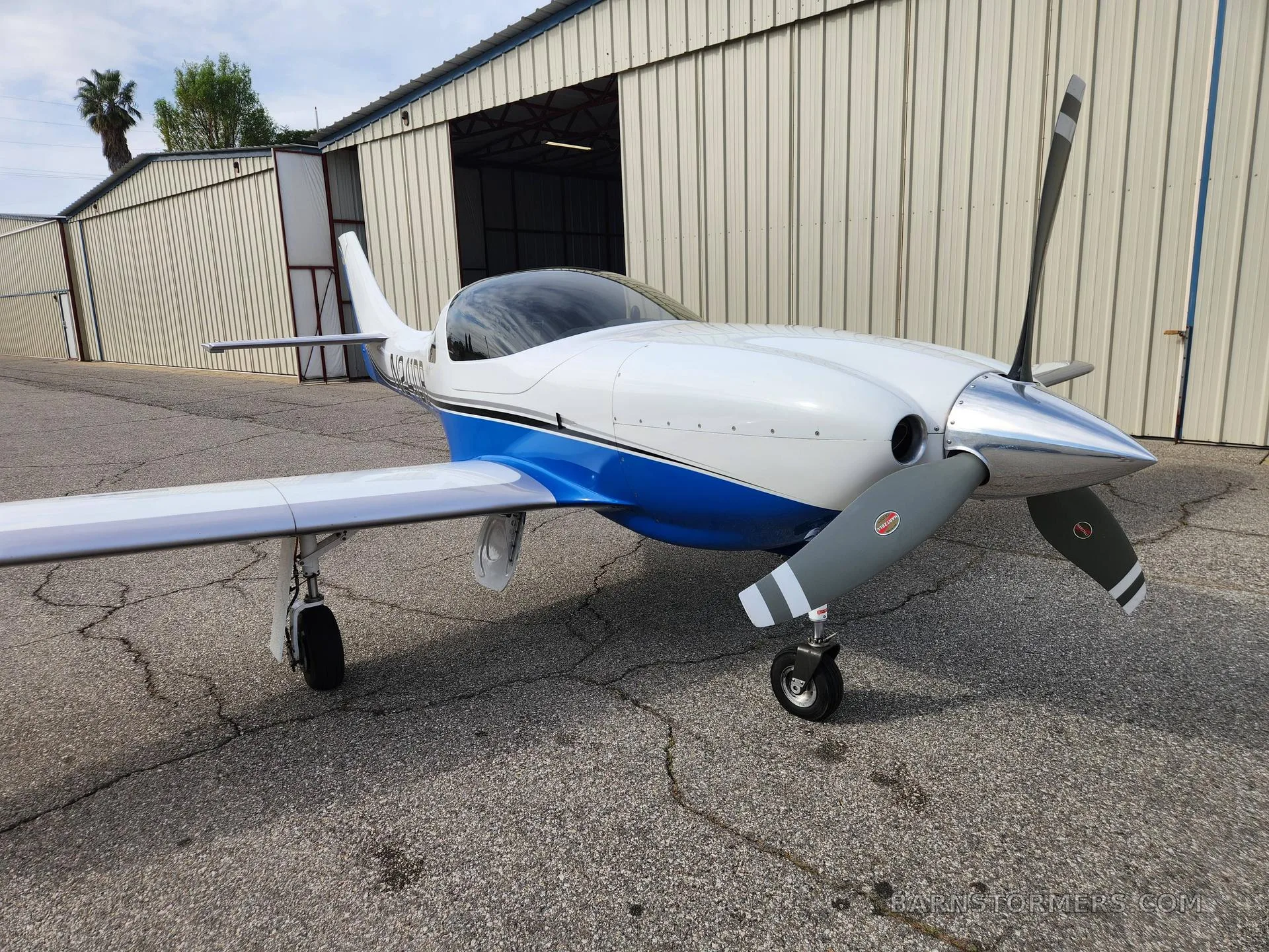 A white and blue single-engine aircraft with a propeller, parked on a tarmac in front of a hangar.