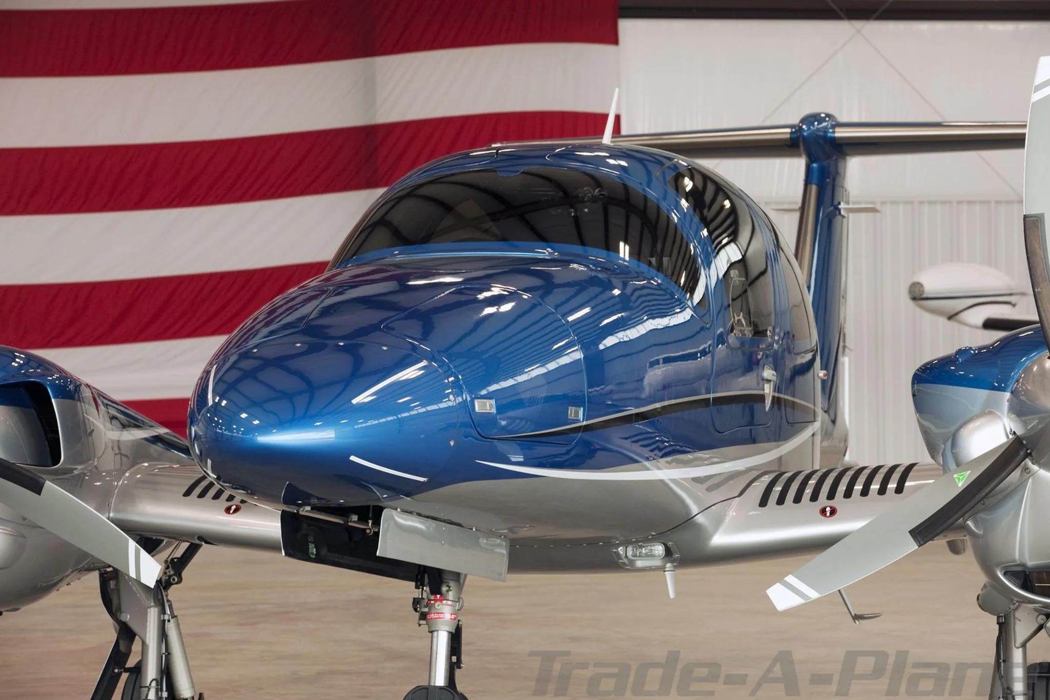 A close-up view of the nose and cockpit of a blue and silver Diamond DA62 aircraft in a hangar.