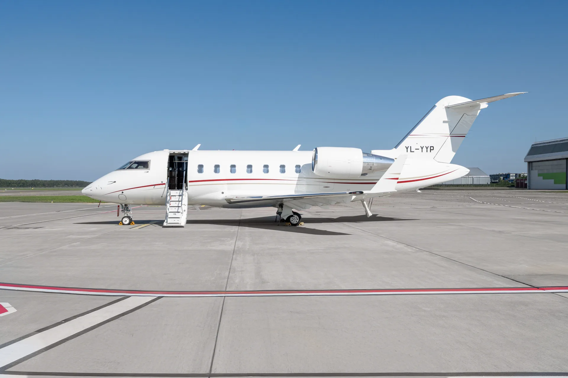 A white Bombardier Challenger 605 business jet with red and black stripes, registration YL-YYP, parked on a tarmac with its door open and stairs extended.