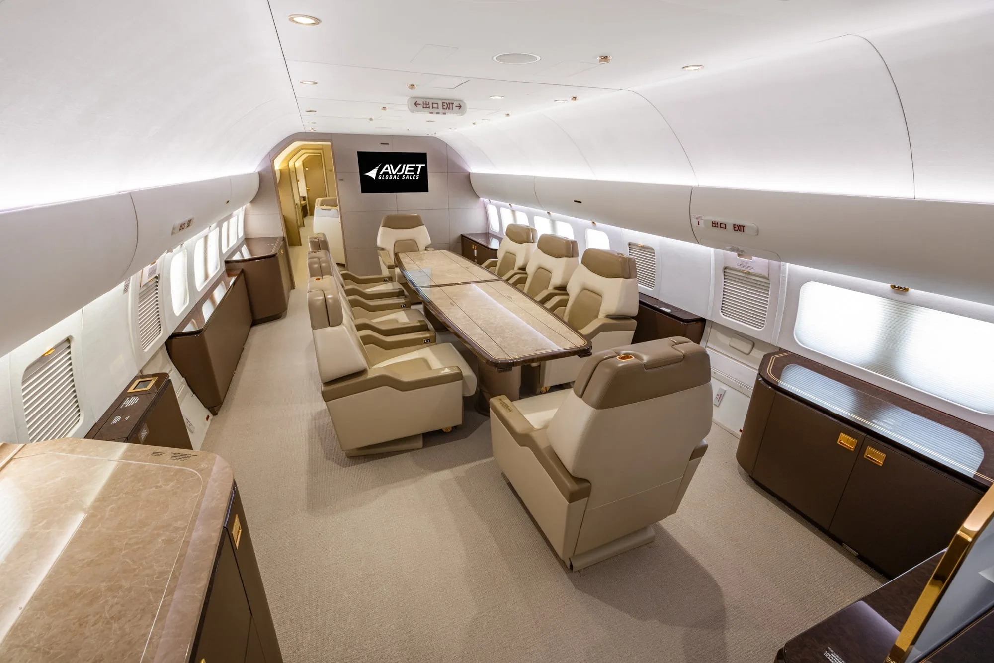 The elegant interior of a private jet cabin with a long dining table surrounded by individual seats and a screen displaying the Avjet logo.