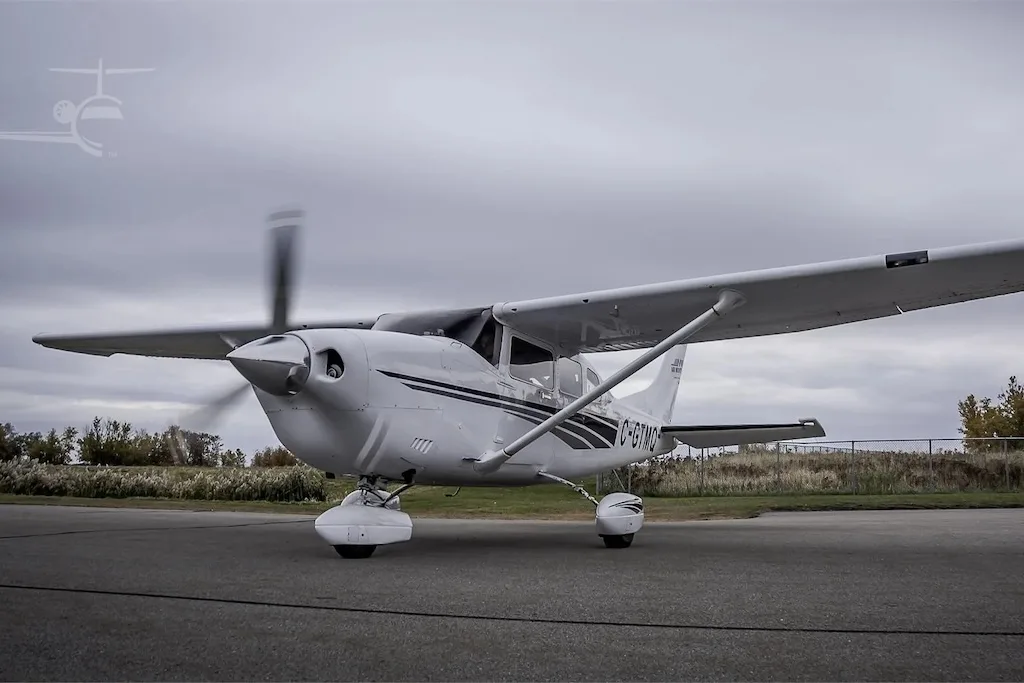 A white single-engine propeller aircraft, registration C-GTMQ, with a spinning propeller, parked on an asphalt runway under an overcast sky.
