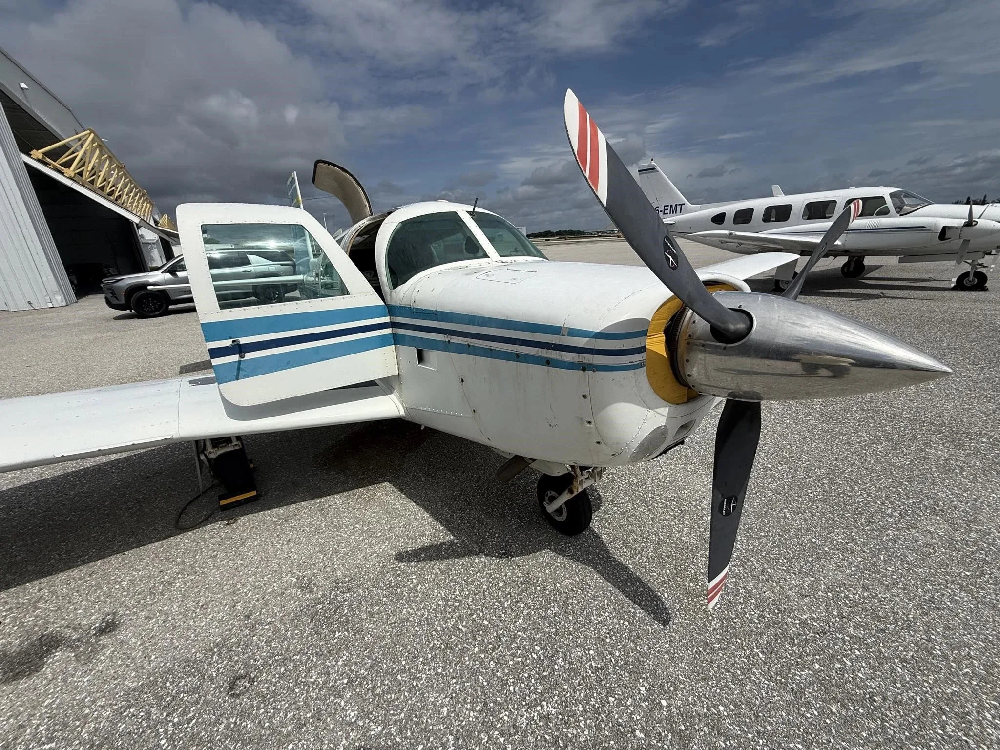 A close-up view of the front-left side of the white single-engine aircraft N9234M with its door open, showing the propeller and blue stripe details.