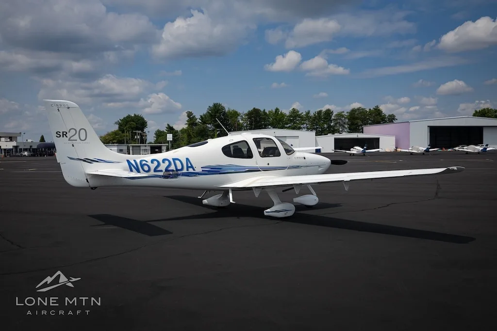 A white Cirrus SR20 aircraft with blue accents and tail number N622DA parked on an airfield with hangars in the background.